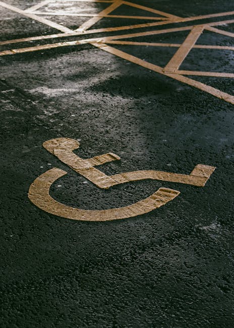 A close-up view of a parking lot surface showing a white-painted disabled parking symbol on dark asphalt. Part of a larger parking area with yellow and white lines indicating designated spaces is visi