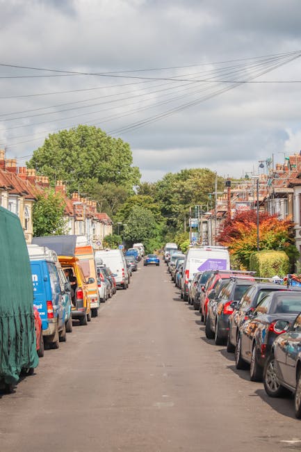 A residential street in Primrose Hill lined with parked cars on both sides, including various makes and models, some covered with protective sheets or tarps. In the background, several houses with bri