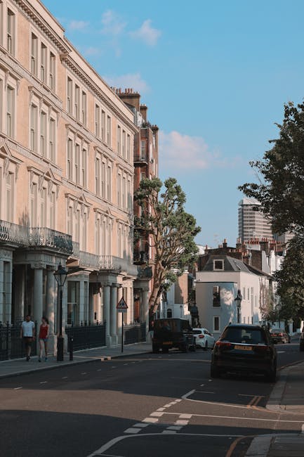 This image depicts a street scene in a residential area, featuring a row of multi-storey Victorian-style buildings with detailed facades and balconies. The buildings are light-colored, and some have black iron railings on the balconies. In the foreground, a few pedestrians are walking along the pavement, and a tree with sparse foliage is situated next to the buildings, providing some greenery. Parked cars are aligned along the curb, including a black vehicle and a white van. The street appears quiet with minimal traffic, and the sky is partly cloudy with patches of blue visible. The environment indicates an urban setting suitable for home relocation or moving services, with the scene showing relevant elements such as vehicles and residential buildings, often associated with house removals and furniture transport. Man and Van Primrose Hill specializes in relocation services within this area, facilitating packing, loading, and transport processes.