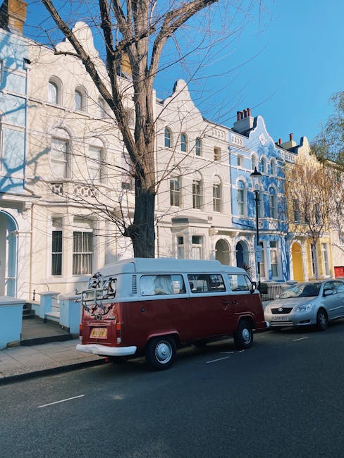 A wide, open park area during daytime featuring a lush green lawn with numerous groups of people sitting on blankets and folding chairs, some engaging in conversation or relaxing. Several trees border the park, providing shade, with others scattered across the area. In the background, a city skyline is visible with multiple high-rise buildings and a prominent telecommunications tower extending upward. The sky above is mostly clear with a few small clouds, and the lighting suggests a bright, sunny day. The scene captures a typical urban park environment where visitors relax outdoors, possibly during leisure or a community event related to house moves or local services, with [COMPANY_NAME] providing household removal and transportation services in NW1, Primrose Hill.